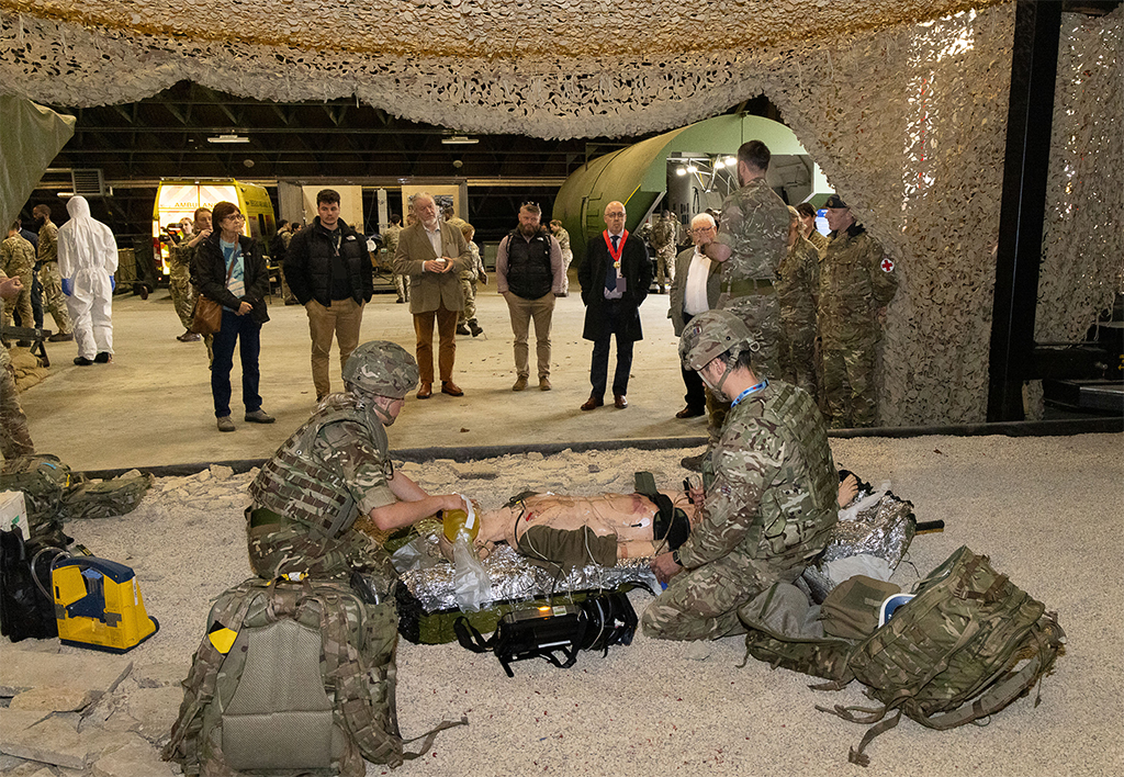 A Tactical Medical Wing team attend to the needs of a training patient in a mock-up of a 'in-theatre' location, while event attendees watch.